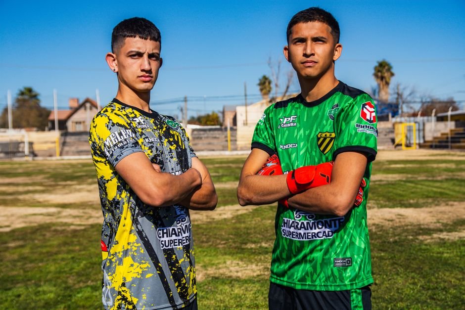 Two young men proudly posing on a soccer field, wearing team jerseys.