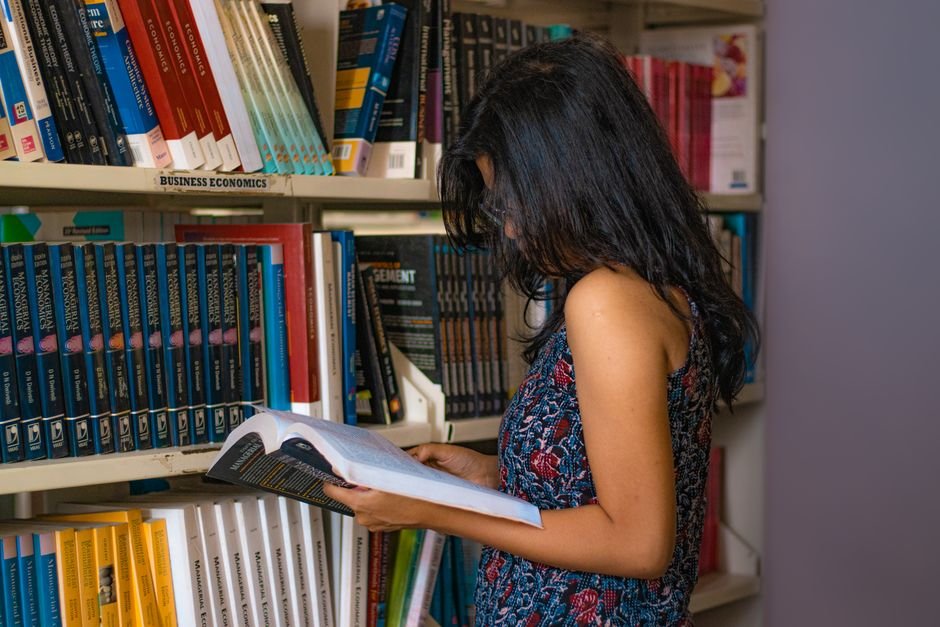 Delhi University Records Highest-Ever UG Registrations for 2026-27 Session Young woman reading a book in a library in Delhi, India, surrounded by shelves of business economics books.