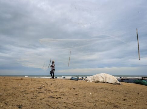 A fisherman skillfully casts a net on the sandy beach of Odisha, India under a cloudy sky.