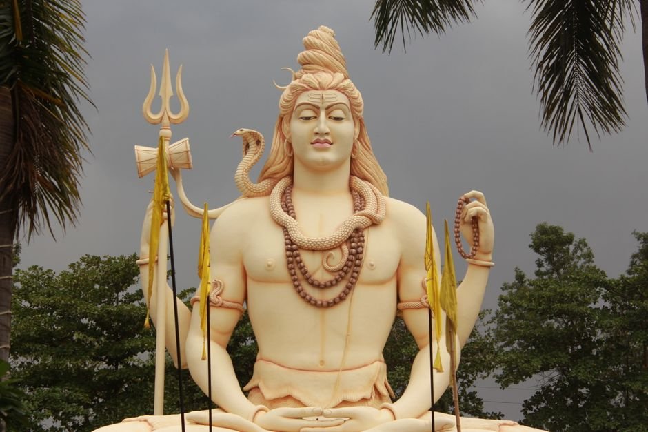 Uttar Pradesh: Devotees Flock to Etawah’s Kedareswar Mahadev Temple During Sawan Month A grand Shiva statue amidst palm trees with a dramatic cloudy backdrop.