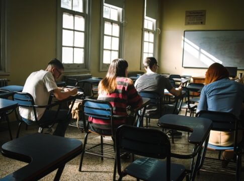 A group of students focused on writing in a sunlit classroom with desks and windows.