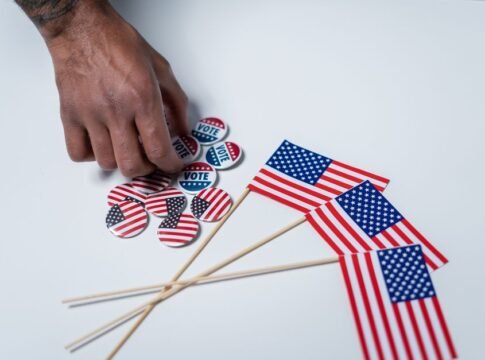 A hand reaches for voting buttons and American flags on a white background.
