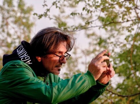 A man in a green jacket capturing nature on his smartphone in Tirana, Albania.