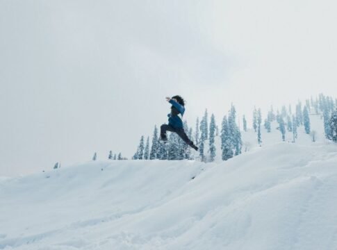 A man leaps across a snow-covered hillside against a backdrop of trees in Gulmarg.