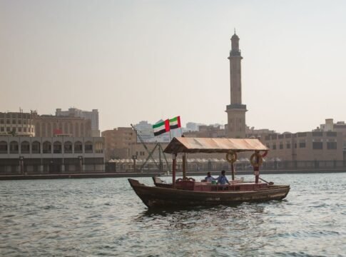 A traditional abra boat sails on Dubai Creek with a city skyline and minaret in the background. A traditional abra boat sails on Dubai Creek with a city skyline and minaret in the background.