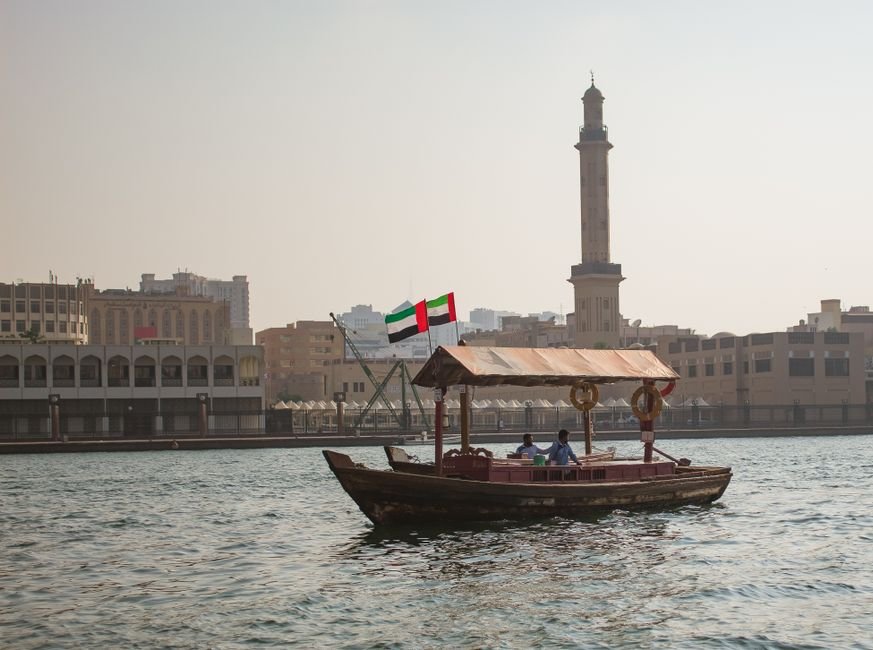 UAE Halts Trade with Iran Following Alleged Missile Launch A traditional abra boat sails on Dubai Creek with a city skyline and minaret in the background.