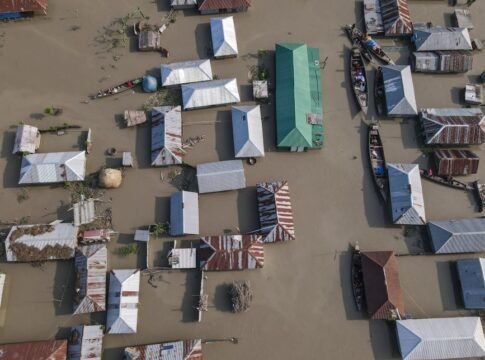 Aerial perspective of a village submerged in floodwaters, highlighting tin-roofed houses and boats.