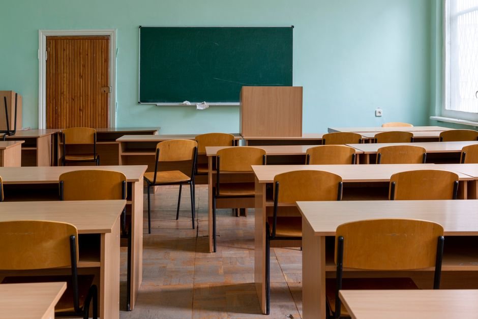 Punjab: Lone Paralyzed Teacher Manages 8 Classes, 25 Students at Selkiana School An empty classroom featuring wooden desks, chairs, and a large green chalkboard.