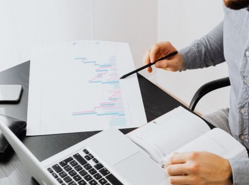 An office setup with a person analyzing a graph printout while using a laptop and notebook.