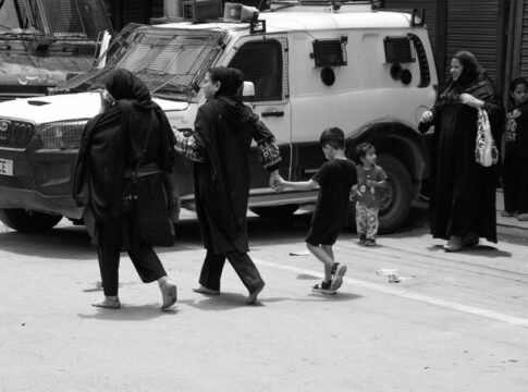 Black and white image capturing people walking past a police vehicle in an urban setting.
