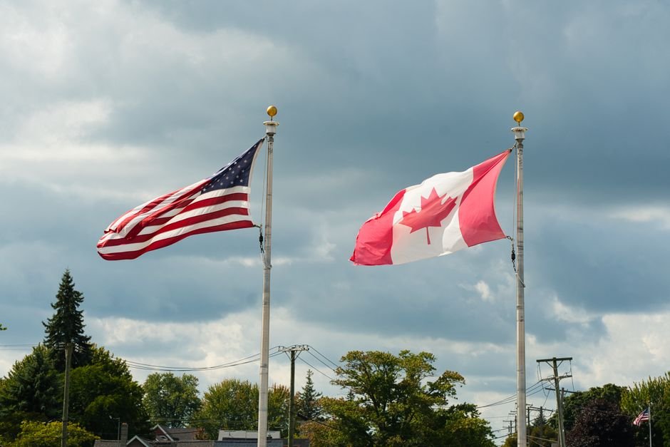 Canada, U.S. in High-Stakes Trade Talks as Tariff Deadline Looms Canadian and American flags waving under a cloudy sky in Harbor Beach, MI.