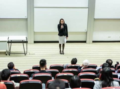 Female speaker presenting to an audience in a modern auditorium setting.