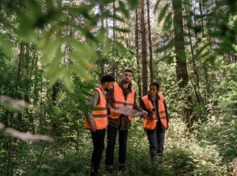 Three adults in safety vests examining a map in a lush forest setting, emphasizing teamwork. Three adults in safety vests examining a map in a lush forest setting, emphasizing teamwork.