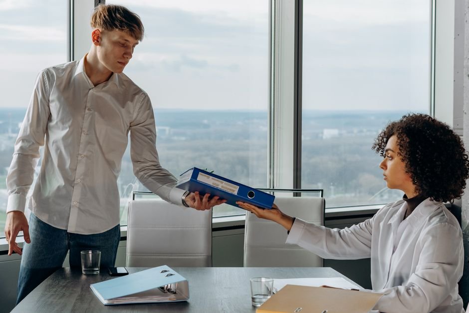 Jammu and Kashmir MBBS Interns Threaten Indefinite Strike Over Low Stipend Two colleagues exchange documents in a contemporary office with a scenic city view.