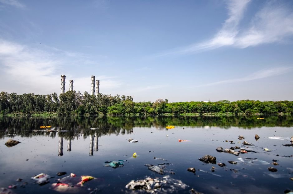 NGT Demands Update on Singhola Waste Accumulation View of a polluted river reflecting factory emissions under a clear sky in India.