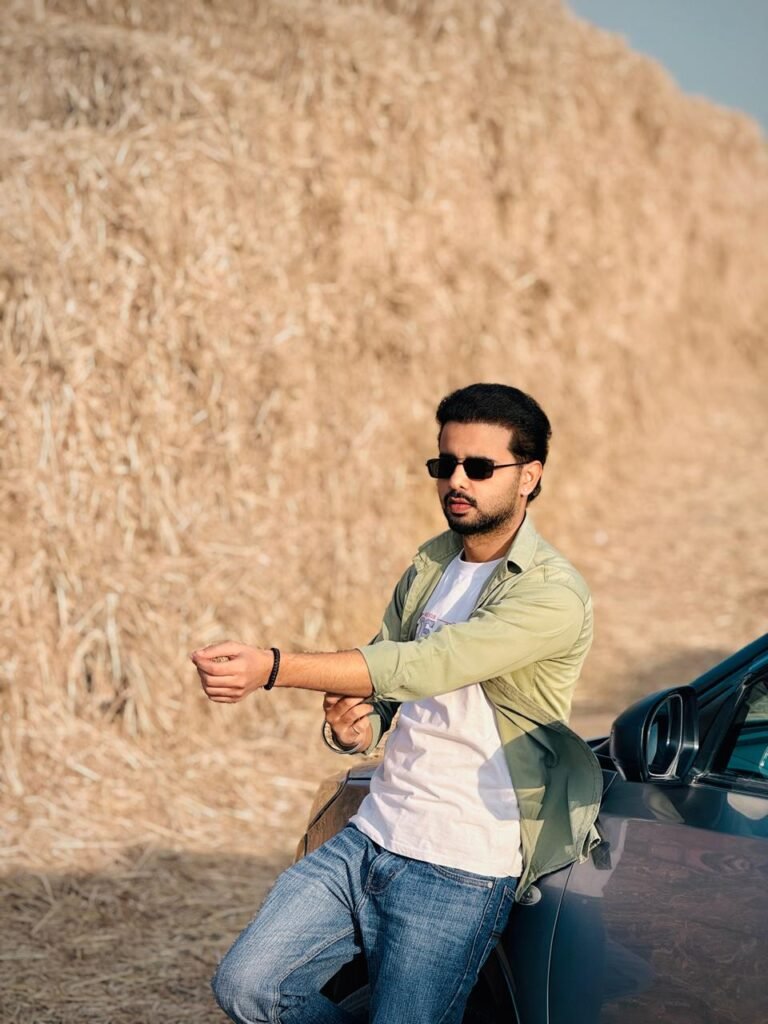 Harinder Cheema leaning against a car in a rural setting with a large stack of hay in the background