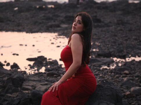 Woman in a red dress sitting on rocks by the beach during sunset, evoking elegance and tranquility.