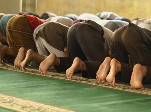 A group of Muslim worshippers praying inside a mosque, captured from behind.