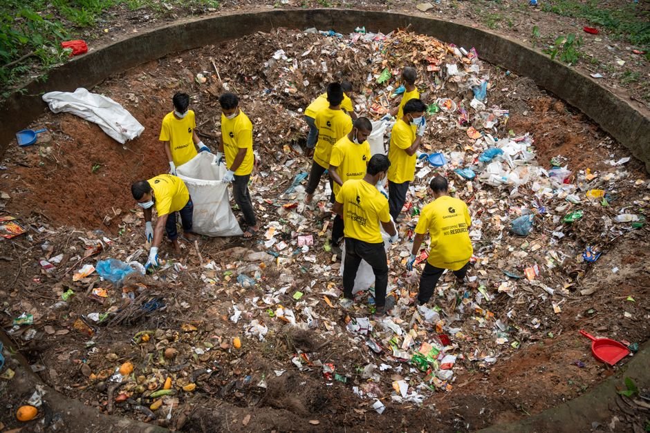 Union Minister Reviews Swachhata Hi Seva 2026 Preparations with States, UTs A group of volunteers in yellow shirts clean up a large waste pit outdoors, demonstrating teamwork and environmental care.