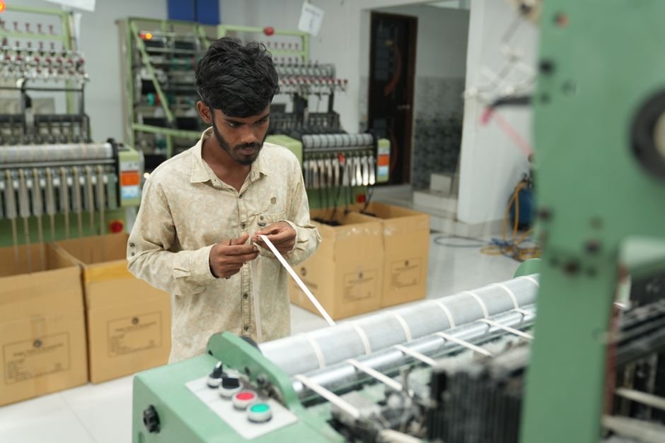 Tiruppur Yarn Producers Urge Garment Units to Modify CCI’s Role Amidst Price Volatility A man focuses on operating a machine in a textile factory, surrounded by large industrial equipment.