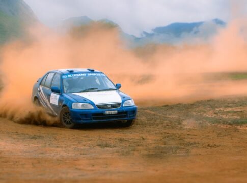 Blue rally car racing on a dirt track with dust clouds.