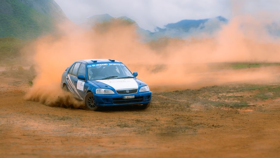 Himachal Rally of Himalayas Navigates Shinkula Pass, Zanskar Valley Blue rally car racing on a dirt track with dust clouds.
