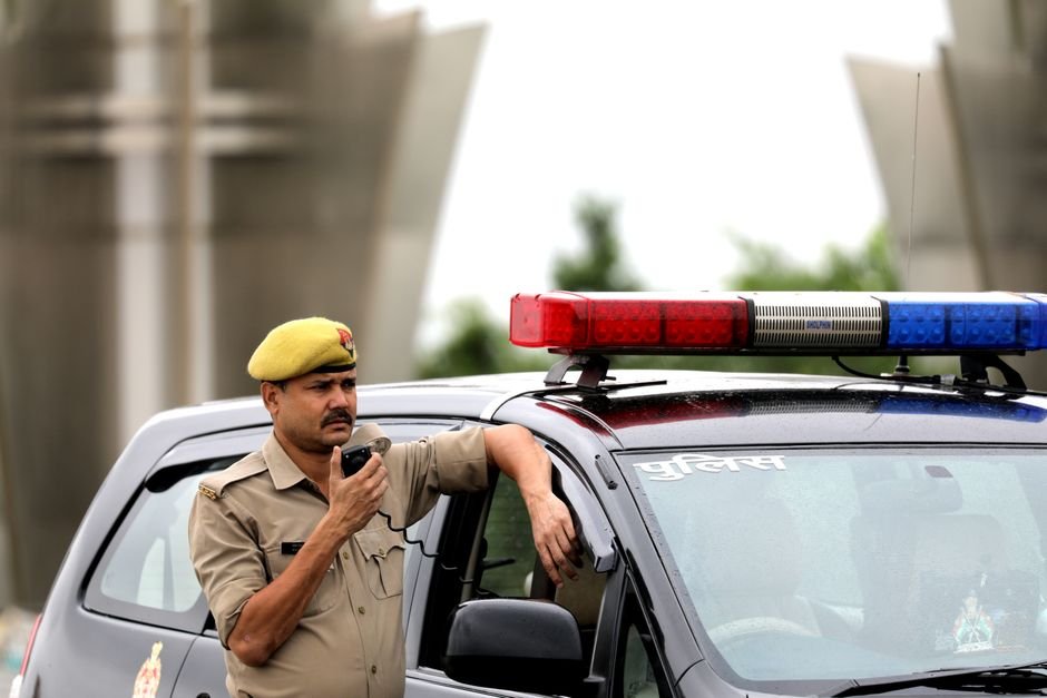 Woman’s Body Discovered in Doda; Identity and Cause of Death Under Investigation Police officer standing beside patrol car holding a radio microphone outdoors.