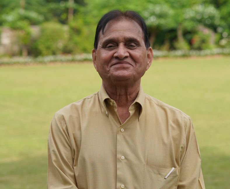 Senior man in a yellow shirt smiling outdoors in a lush green garden.
