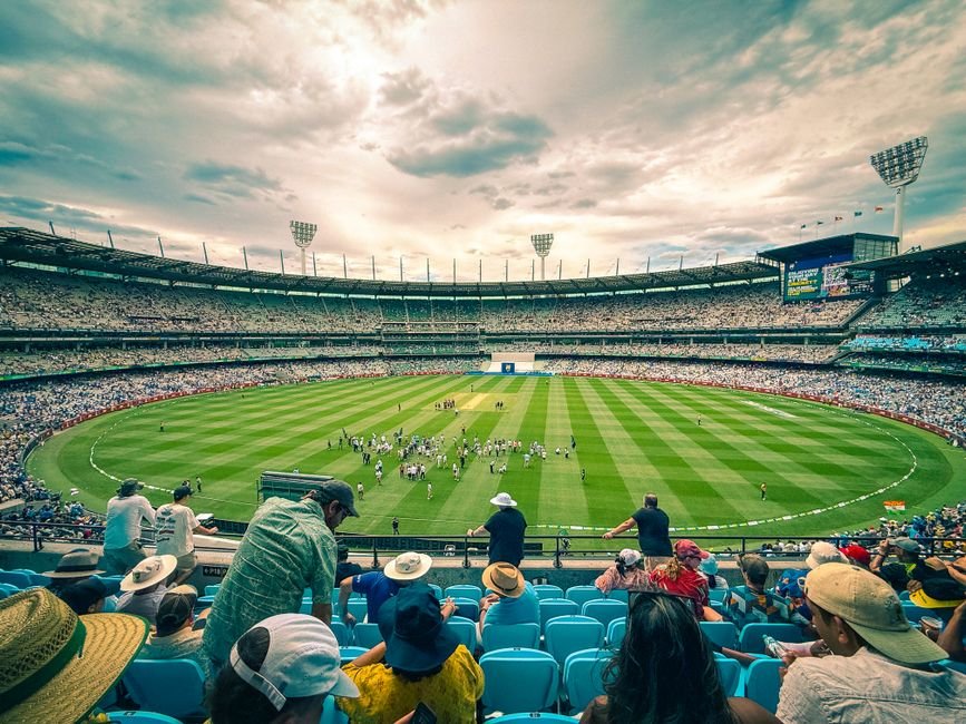 Lucknow Fan Walks 2,390 km to Meet Cricketer Hardik Pandya, Receives Hug and Ticket Home A lively cricket match at Melbourne Cricket Ground with a colorful crowd under a dynamic sky.