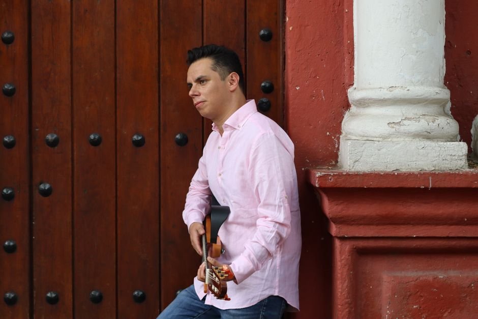 J. A. Bayona, Javier Bardem Join Forces for Documentary on Cinematographer Néstor Almendros A musician in a pink shirt plays guitar by a rustic wooden doorway, creating a serene street scene.