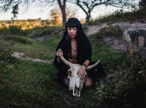 A woman in a black hooded cloak holding a large animal skull in a grassy outdoor setting. A woman in a black hooded cloak holding a large animal skull in a grassy outdoor setting.
