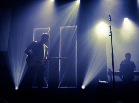 Anonymous male musician performing melody with guitar on stage near microphone and DJ in dark club with spotlights