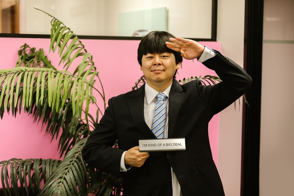 Jimmy Fallon and Jamie Lee Curtis Parody Tech Moguls in ‘You Can See Everything’ Skit Asian man saluting in formal attire, holding a humorous sign indoors.
