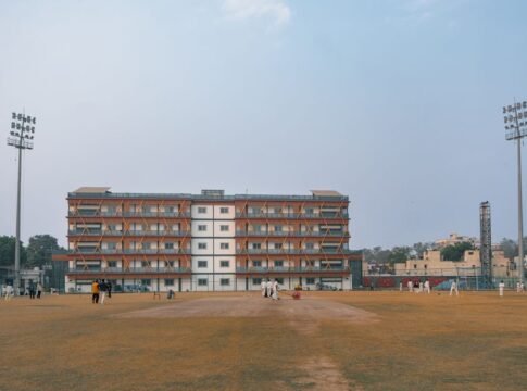 Daytime cricket match on a large sports field in Delhi, India with players and floodlights. Daytime cricket match on a large sports field in Delhi, India with players and floodlights.