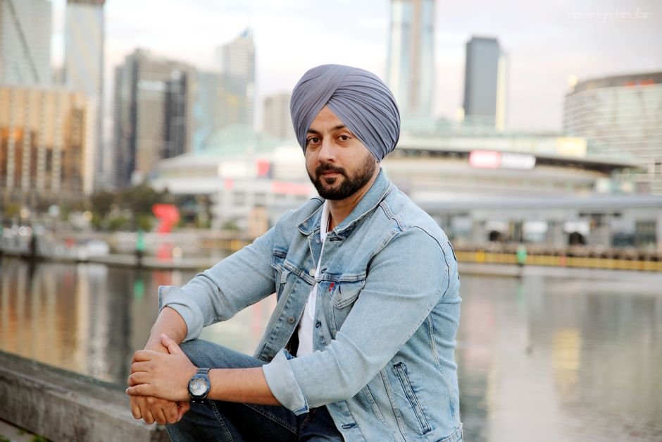 Punjab Congress Leader Simarjeet Bains to Challenge Defamation Sentence in Higher Courts Man wearing denim jacket and turban sitting by the waterfront in a cityscape backdrop.