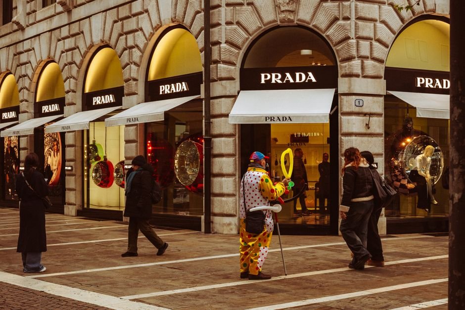 Prada Taps Disney Animator John Pomeroy for Hand-Drawn Holiday Campaign Starring Sadie Sink and Maya Hawke People passing by a vibrant clown entertainer outside a Prada store in Padova, Italy.