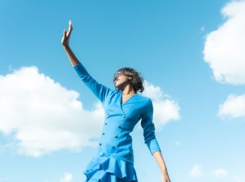 Stylish woman in blue dress reaching up, set against a bright blue sky.