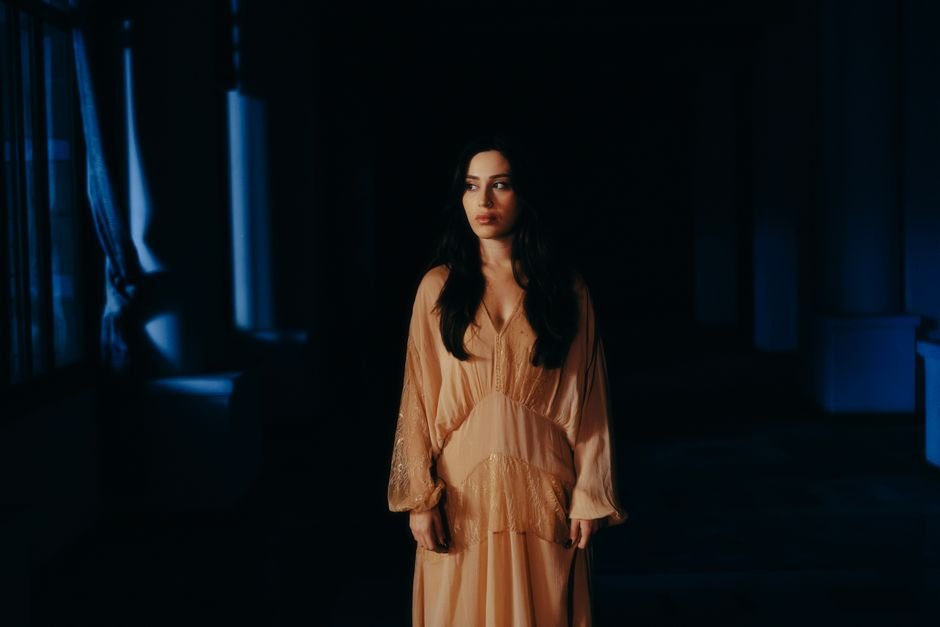 Kacey Musgraves Expands ‘Middle of Nowhere’ Universe with New Deluxe Tracks Woman in a flowing dress stands by a window in dramatic lighting.
